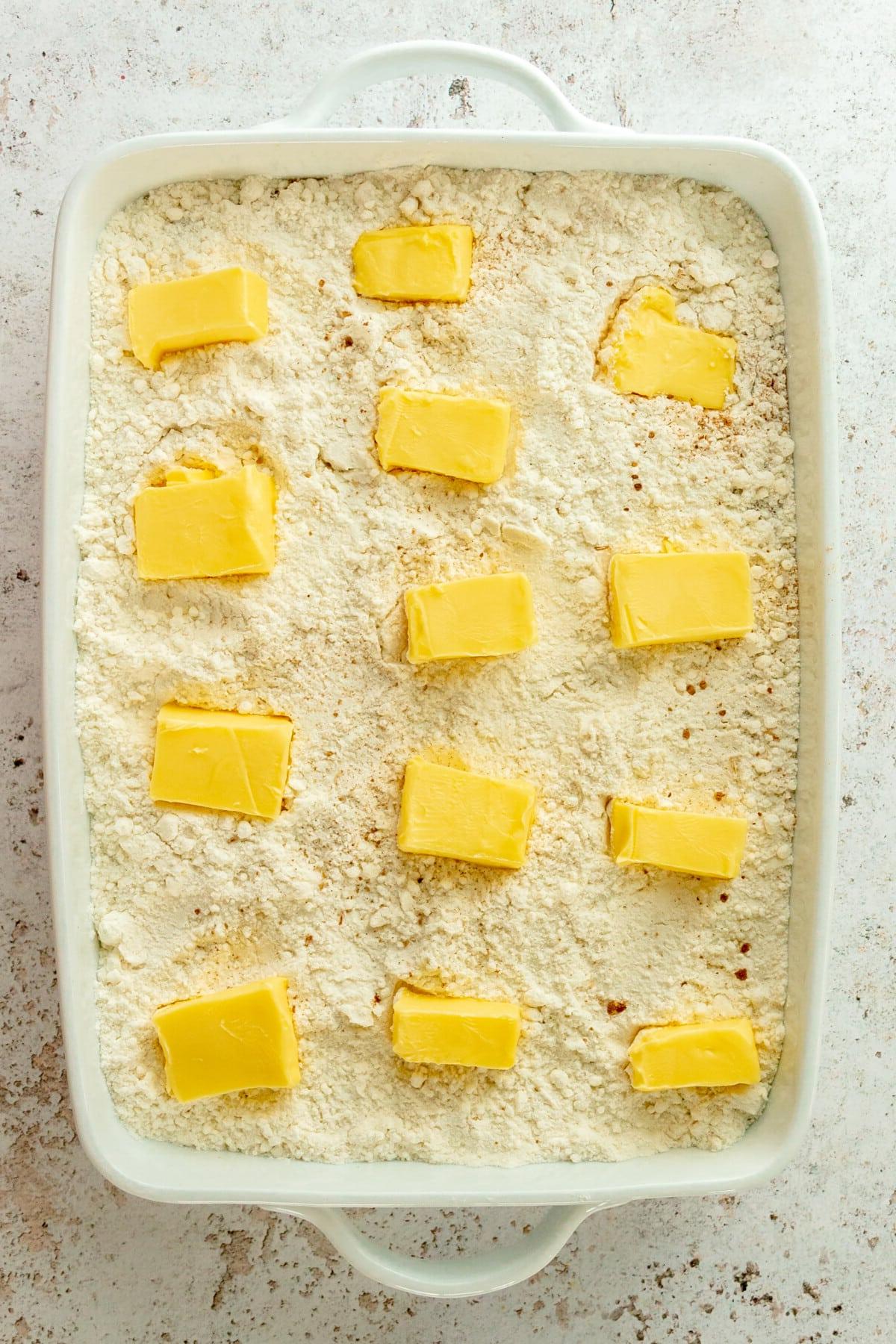 Kürbis-Dump-Kuchen 6 Butterscheiben liegen auf einer Kürbis-Dump-Cake-Mischung in einer weißen, rechteckigen Auflaufform auf hellgrauem Untergrund.