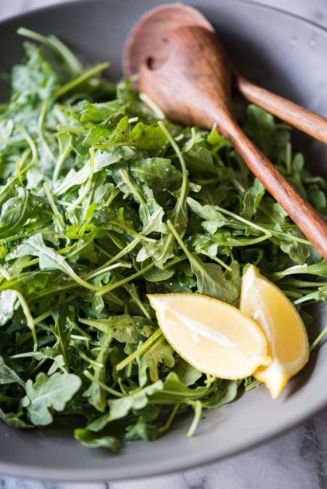 Rucola Salat mit Zitrone und Meersalz 6 Rucola Salat in einer grauen Schüssel mit zwei Zitronenspalten und Holzbesteck