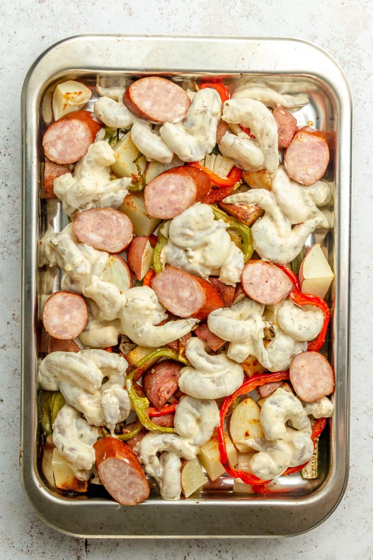 Cajun Garnelen vom Backblech 7 Mit Remouladensoße überzogene Garnelen und Andouille-Wurststücke liegen auf geröstetem Gemüse auf einem Edelstahl-Backblech mit Rand auf einer hellgrauen Oberfläche.