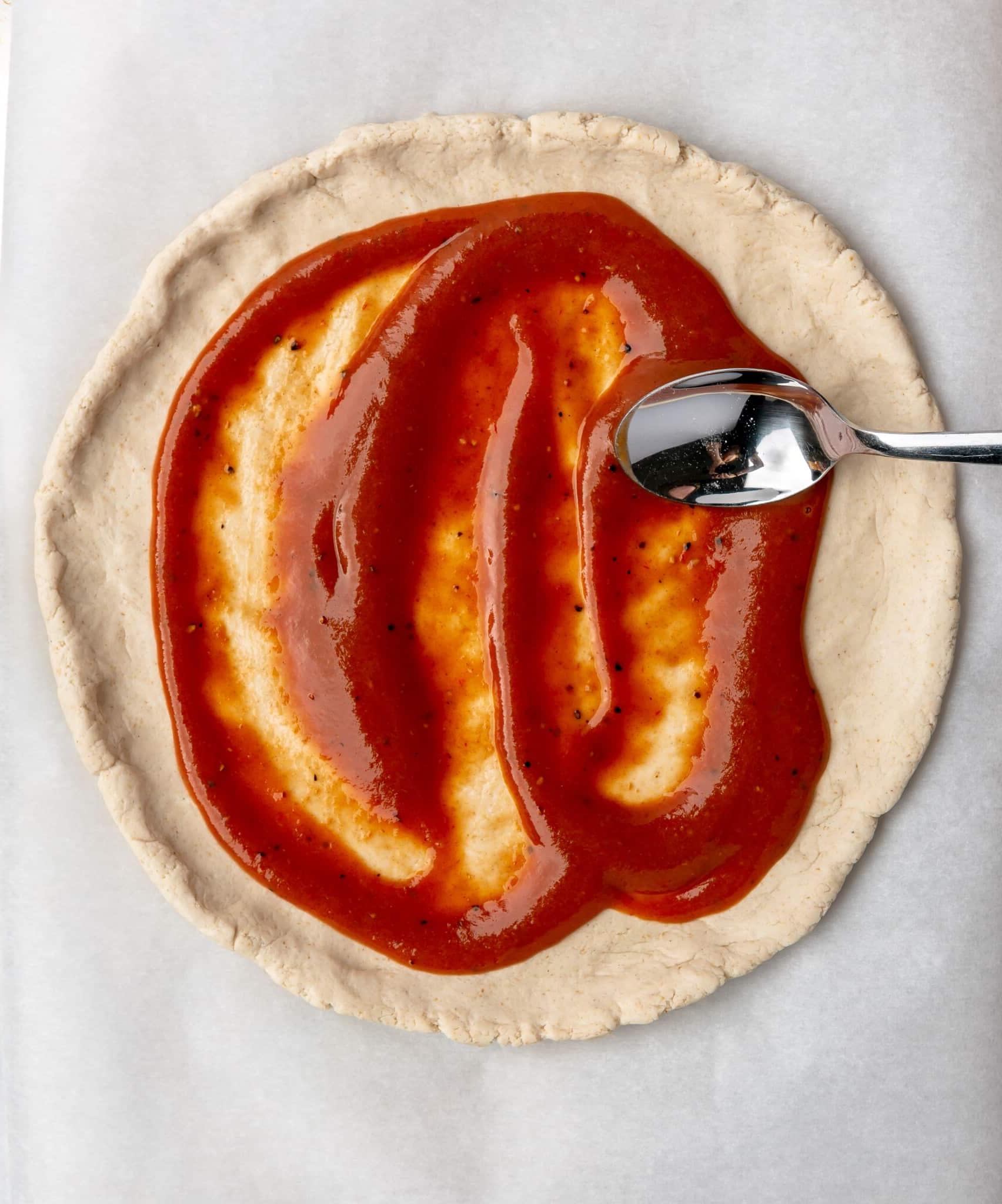 Eine Person, die mit einem Löffel BBQ-Sauce auf Pizzateig verteilt.