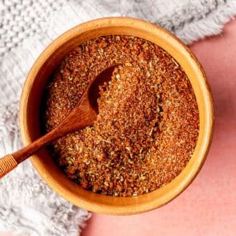 Beste Trockenmarinade für Schweinefilet 5 Draufsicht auf eine braune Tonschale mit einem kleinen Holzlöffel, gefüllt mit einer Gewürzmischung. Die Schale steht auf einer rosa Oberfläche mit einem hellgrauen, strukturierten Leinen.