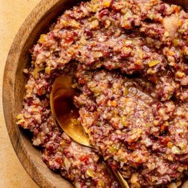 Oliven-Tapenade 7 Oliven-Tapenade in einer hölzernen Servierschale. Ein goldfarbener Löffel liegt daneben.