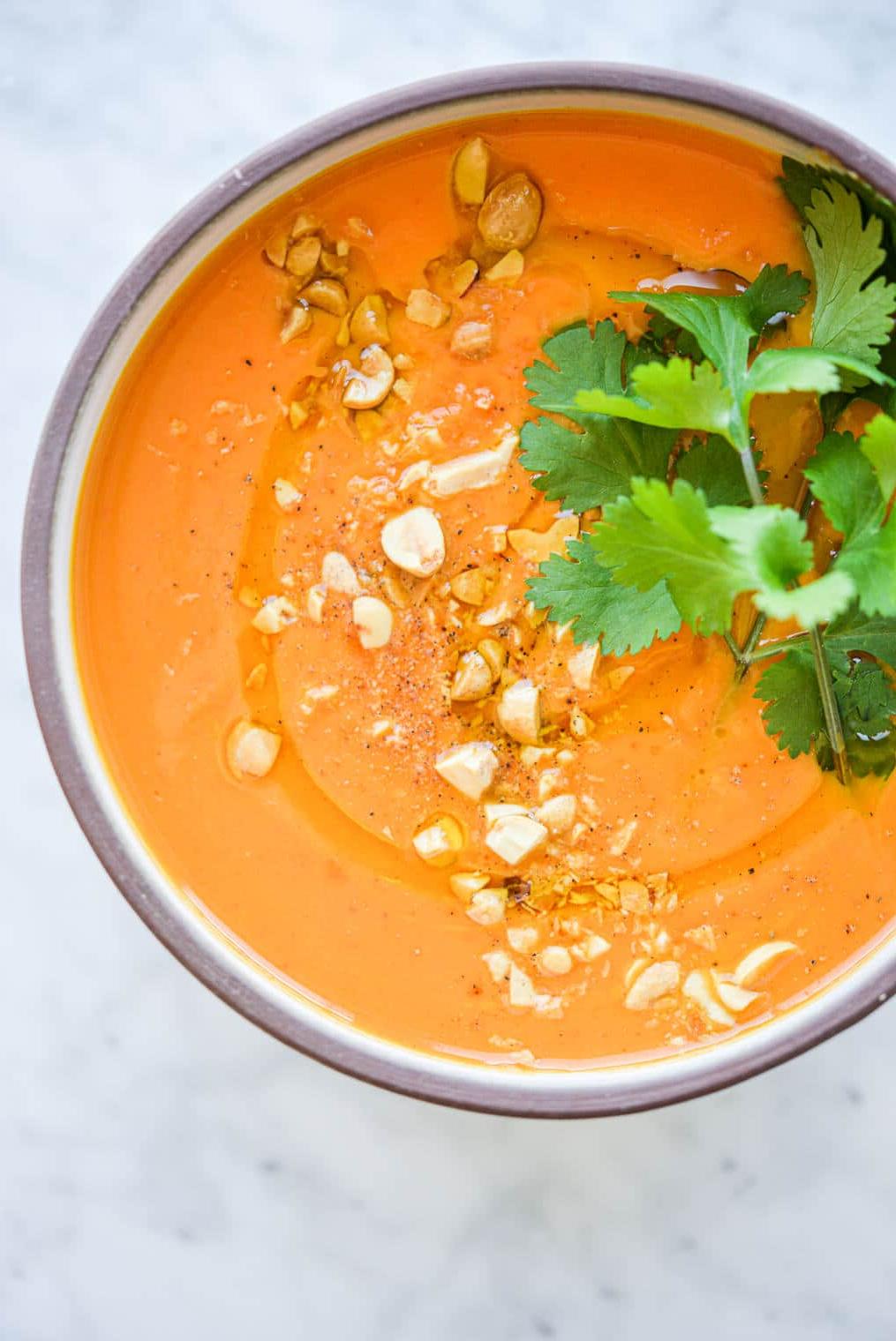 Thailändische Karotten-Ingwer-Suppe 8 Eine Schüssel mit leuchtend oranger Thai-Karottensuppe, garniert mit frischem Koriander und zerdrückten Erdnüssen auf einer Marmoroberfläche.