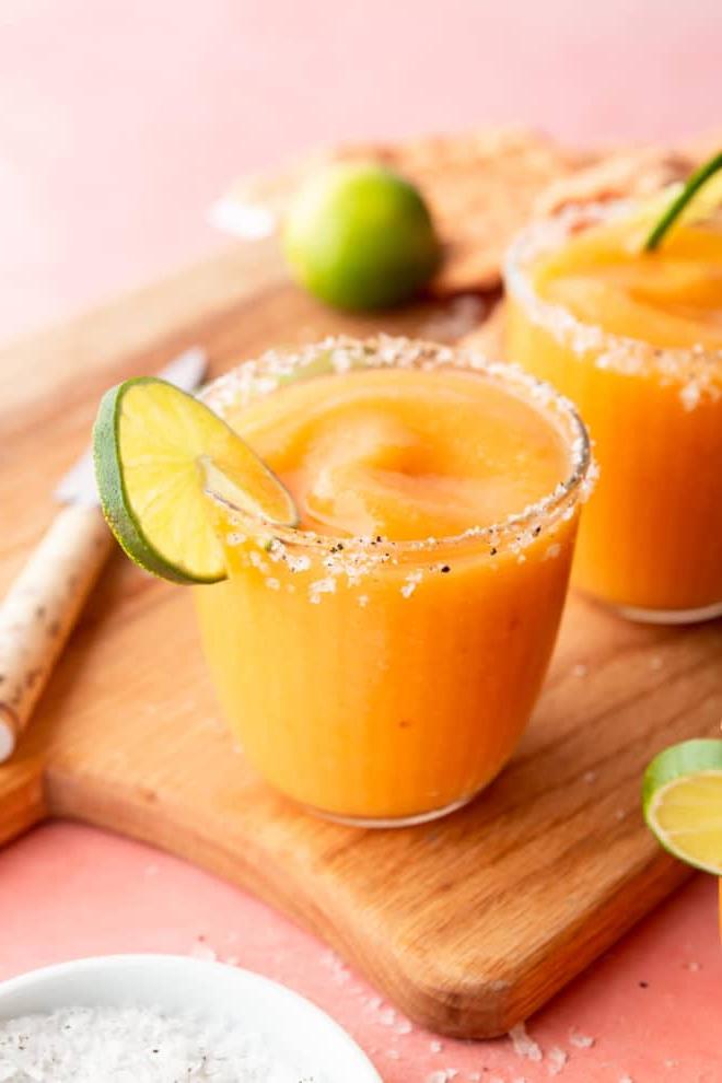 Das beste Pfirsich-Margarita-Rezept 4 Seitenansicht von gefrorenen Pfirsich Margaritas in Gläsern, garniert mit Meersalzflocken und einer Limettenscheibe, auf einer pinkfarbenen Oberfläche mit Schneidebrett, Schälmesser, einer Prise Meersalzflocken, Limettenscheiben und orangefarbenem Leinentuch.