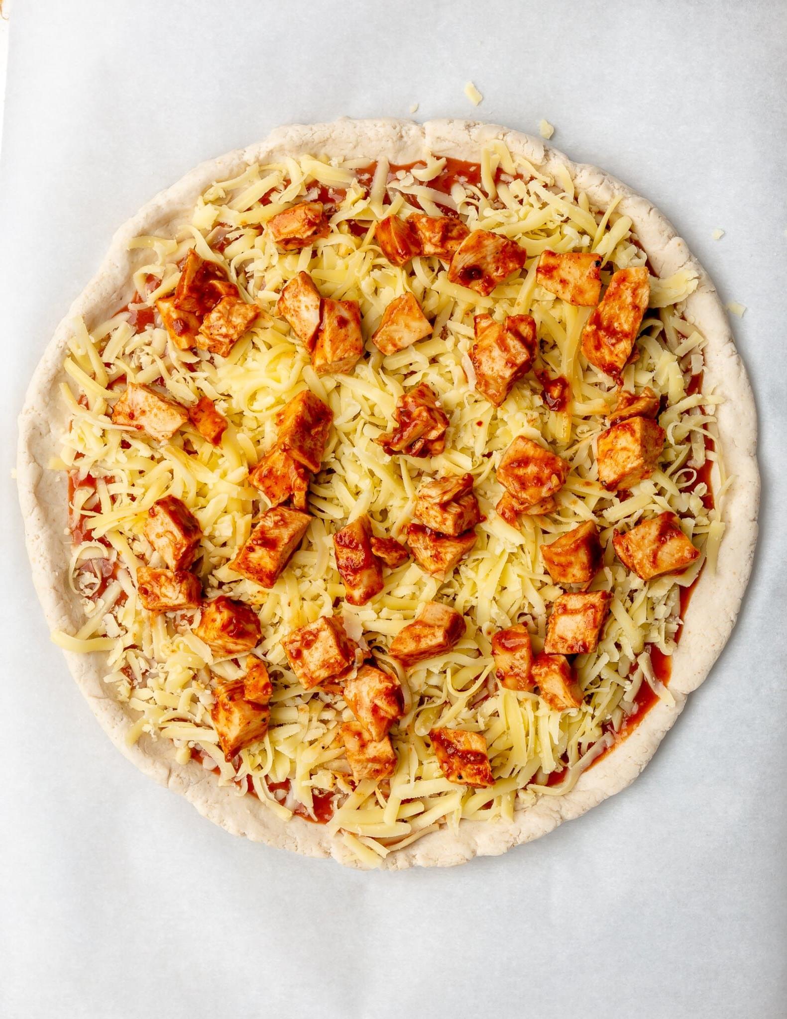 Roher Pizzateig belegt mit BBQ-Sauce, BBQ-Hähnchen und Mozzarella-Käse.