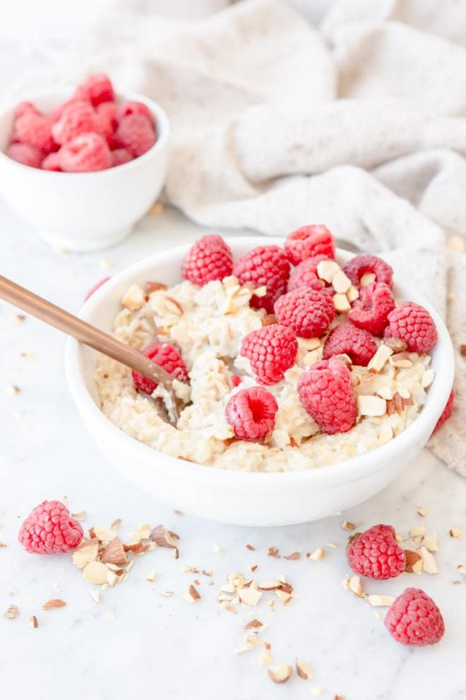 Wie man Haferbrei auf dem Herd zubereitet 4 Oatmeal in a white bowl topped with chopped almonds and raspberries with a small white bowl of raspberries and a beige linen in the background.