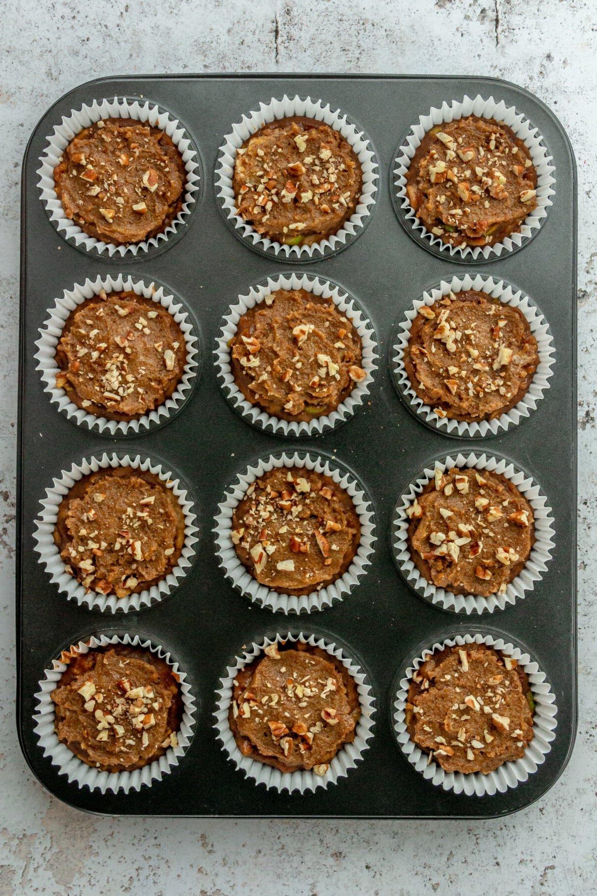 **Paläo Apfel-Zimt-Muffins** 8 Paleo apple cinnamon muffin batter topped with a crumble mixture and pecans batter sits in white liners in a 12 muffin tin on a light grey surface.