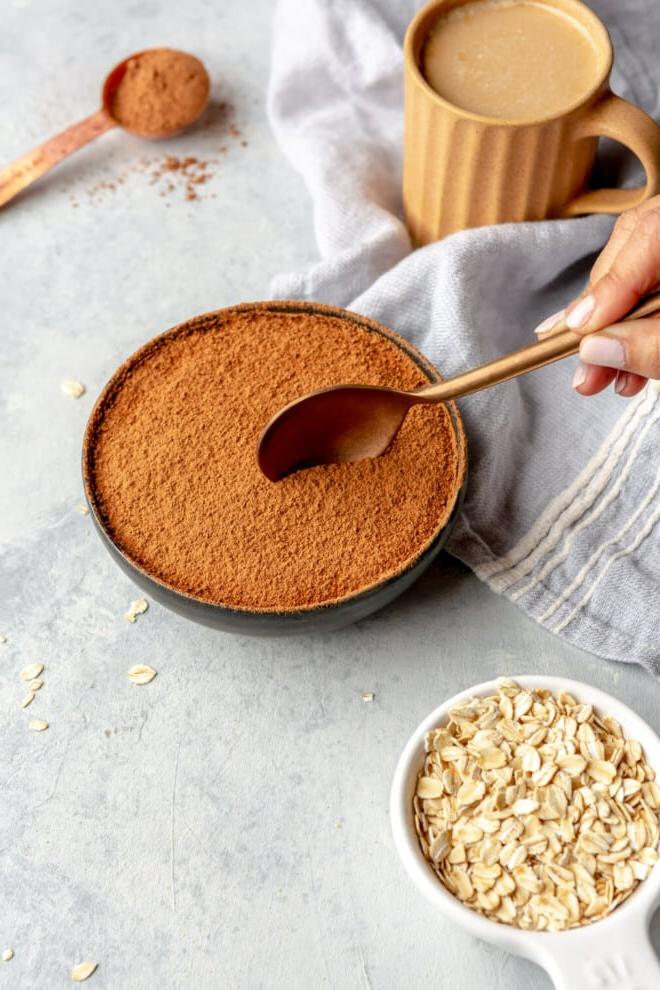 Tiramisu Overnight Oats 4 Hand hält einen Löffel und bereitet sich darauf vor, eine Portion Overnight Oats aus einer Schale zu nehmen. Auf dem Tisch stehen außerdem eine Tasse Kaffee, ein Messlöffel mit Kakaopulver und ein Messbecher mit trockenen Haferflocken.