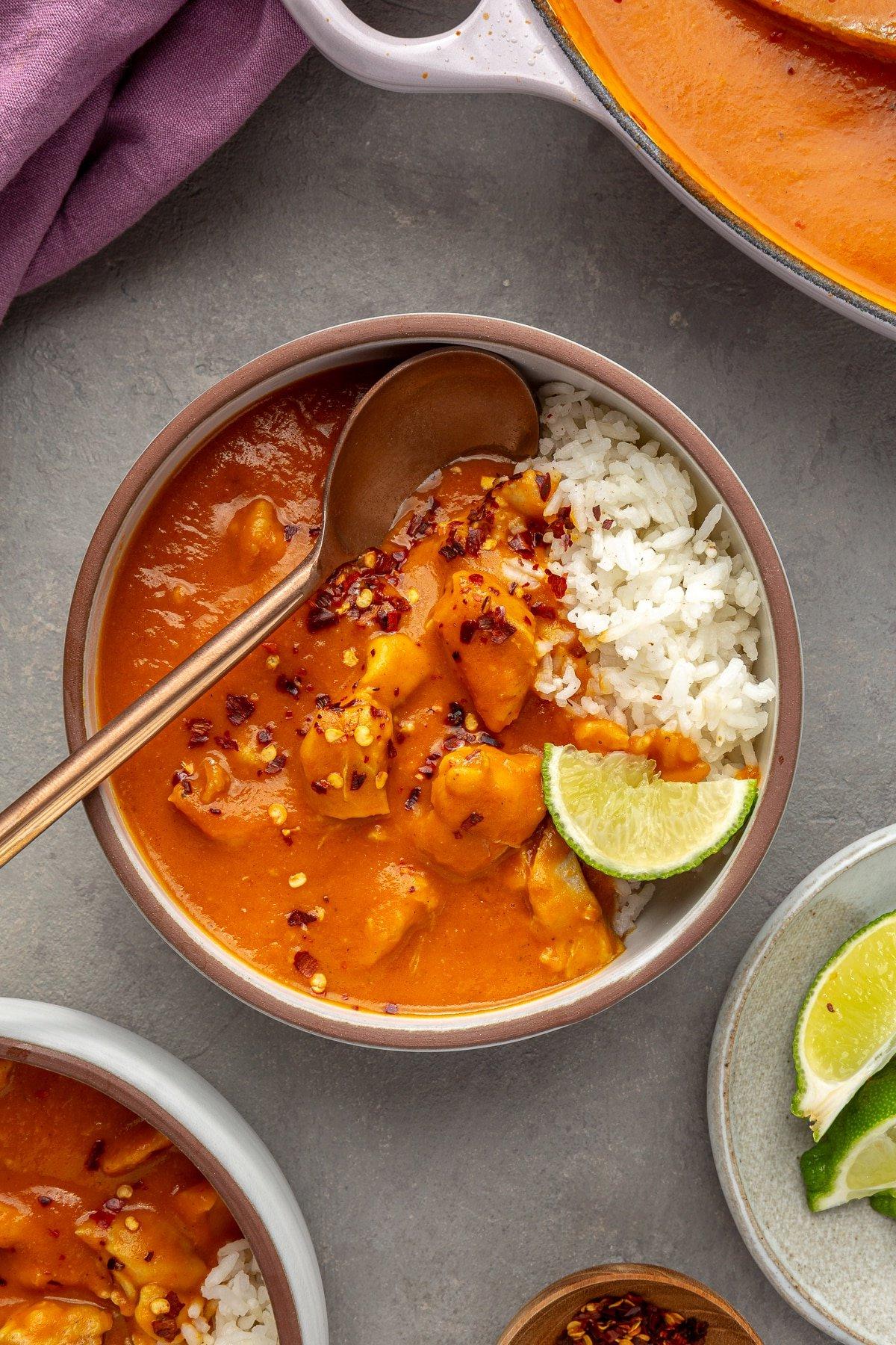 Eine Servierschüssel mit Reis ist mit Kürbis-Curry belegt. Ein Kupferspoon steckt in der Schüssel zusammen mit einer Limettenscheibe.