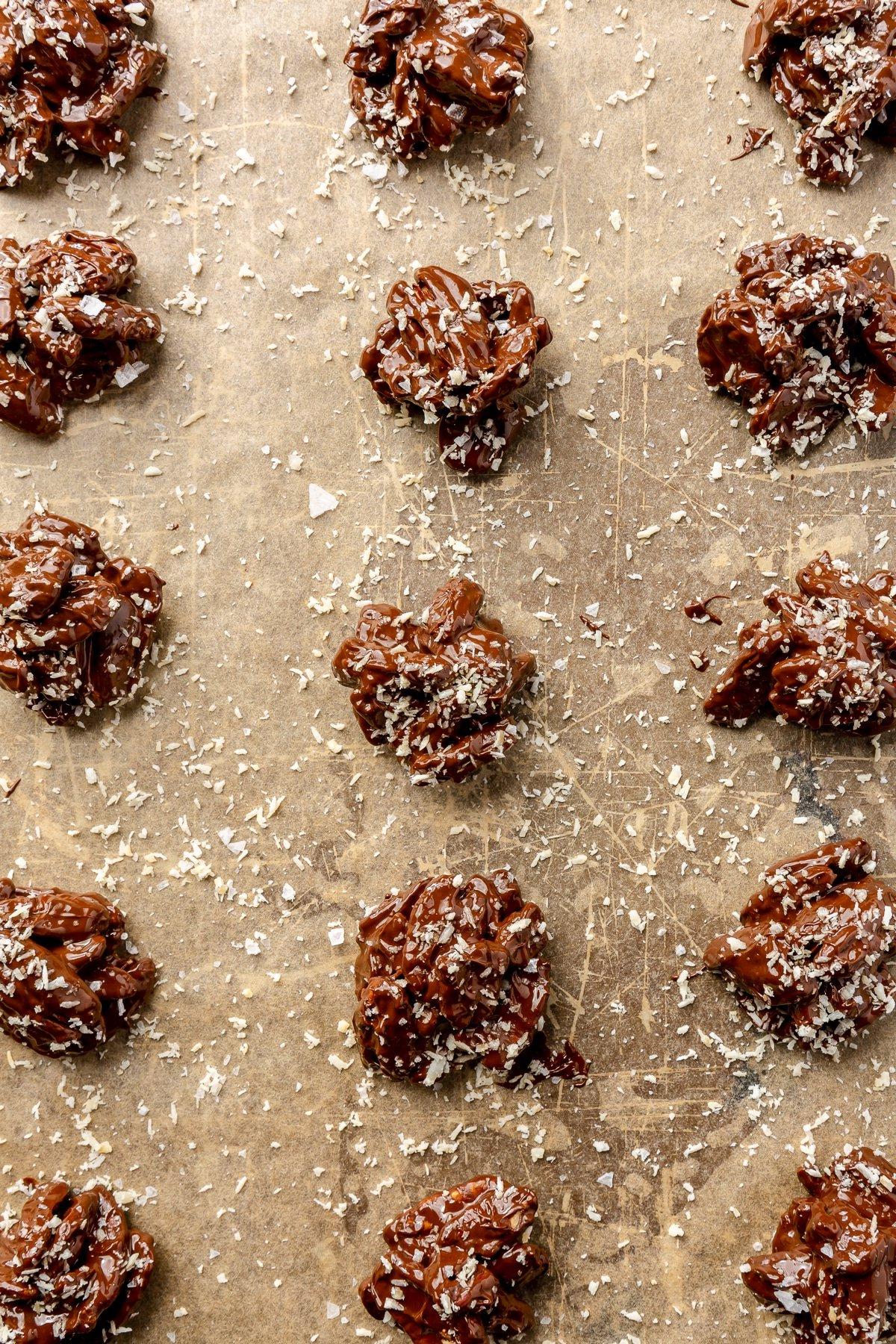 Frisch geformte Schoko-Chili-Pekannuss-Häufchen auf Backpapier, bereit zum Bestreuen mit Kokosraspeln und Meersalz.
