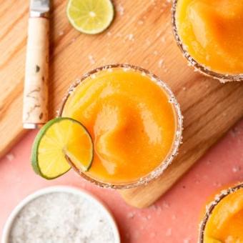 Das beste Pfirsich-Margarita-Rezept 5 Aufnahme von oben: Gefrorene Pfirsich Margaritas in Gläsern, garniert mit Meersalzflocken und einer Limettenscheibe, auf einer pinkfarbenen Oberfläche mit Schneidebrett, Schälmesser, einer Prise Meersalzflocken, Limettenscheiben und orangefarbenem Leinentuch.