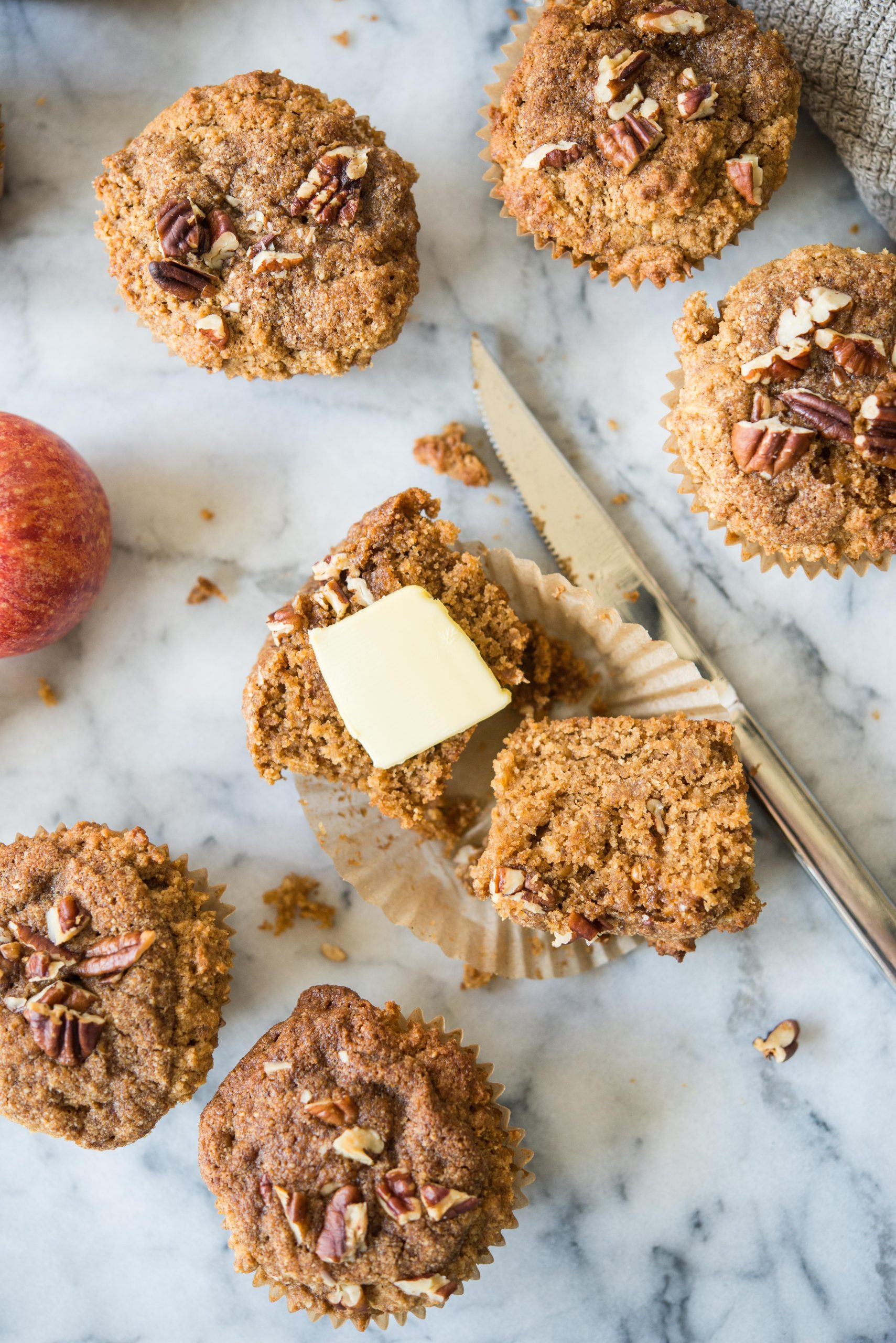 **Paläo Apfel-Zimt-Muffins** 9 an apple cinnamon muffin cut in half with butter on top surrounded by more muffins all on a marble countertop
