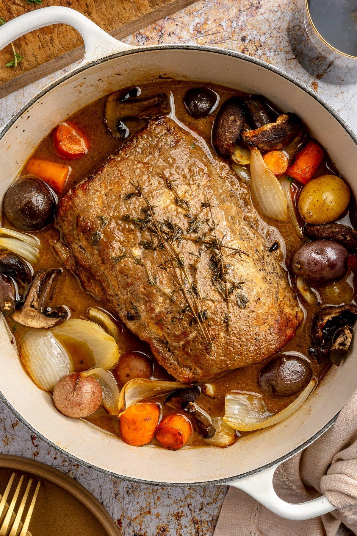 **Balsamico-Schweinelendenrezept** 9 Ein fertig gegarter Balsamico-Schweinebraten liegt in einem weißen, emaillierten Topf. Zwei Weingläser, ein brauner Teller mit goldenem Besteck und ein Schneidebrett stehen daneben.
