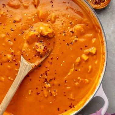 Ein weißer Topf mit Kürbis-Curry, das mit roten Pfefferflocken bestreut ist. Ein Holzlöffel ist zu sehen, wie er einen Teil des Currys schöpft.
