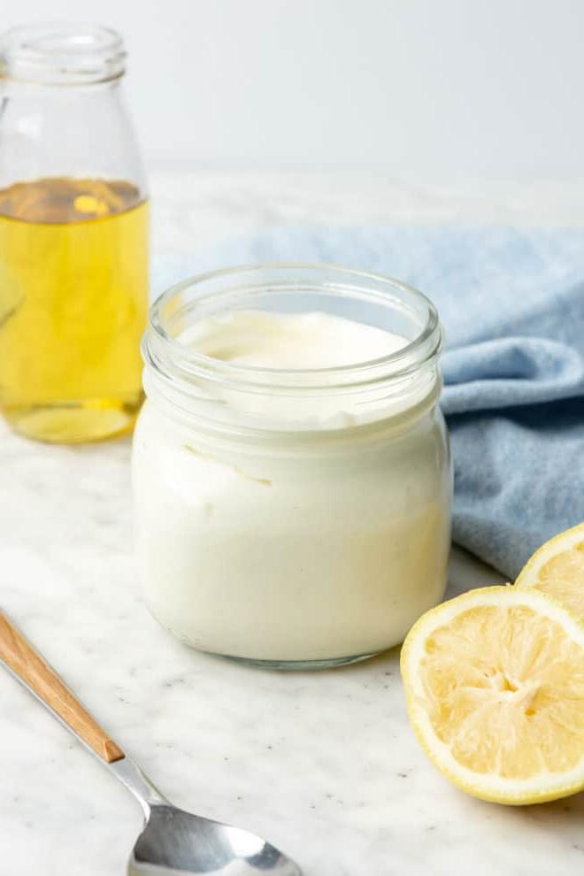 Einfaches Selbstgemachtes Mayonnaise Rezept 4 Glas mit hausgemachter Mayonnaise auf einer grau-weißen Theke mit Zitronenhälften, Avocadoöl im Hintergrund, einem Servierlöffel und einem hellblauen Leinentuch.