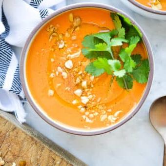 Thailändische Karotten-Ingwer-Suppe 10 Eine Schüssel mit leuchtend oranger Thai-Karottensuppe, garniert mit frischem Koriander und zerdrückten Erdnüssen auf einer Marmoroberfläche.