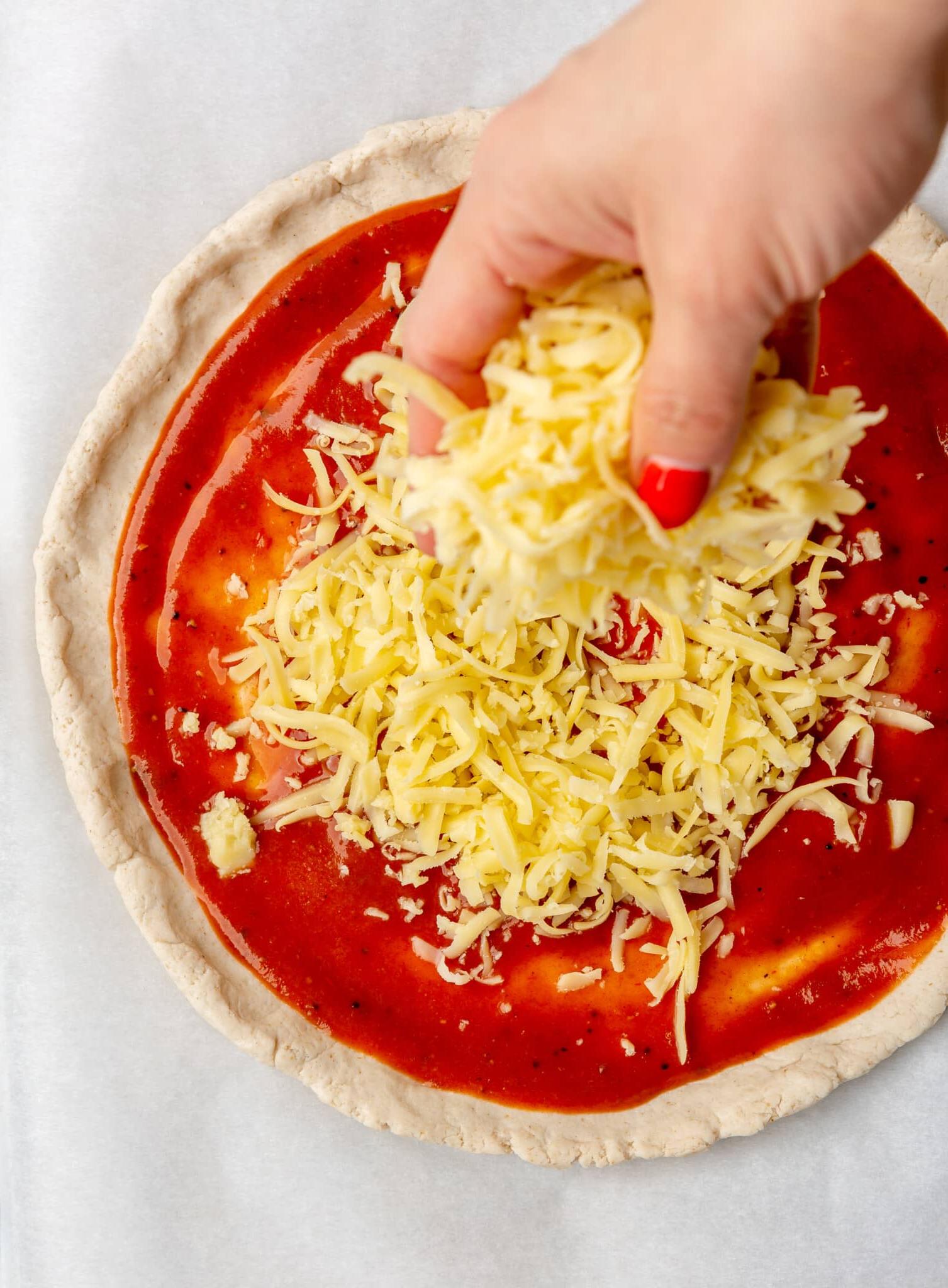 Eine Person, die mit den Händen Mozzarella-Käse auf Pizzateig streut.