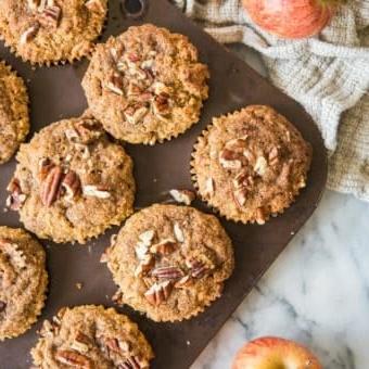 **Paläo Apfel-Zimt-Muffins** 10 paleo apple cinnamon muffins in a muffin tin on a marble board with apples and a grey cloth surrounding them