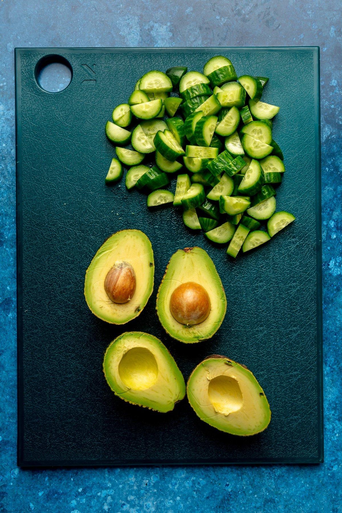 Harissa-Honig-Hähnchen-Bowls 10 Geschnittene Gurken und halbierte Avocados liegen auf einem dunkelblauen Schneidebrett.