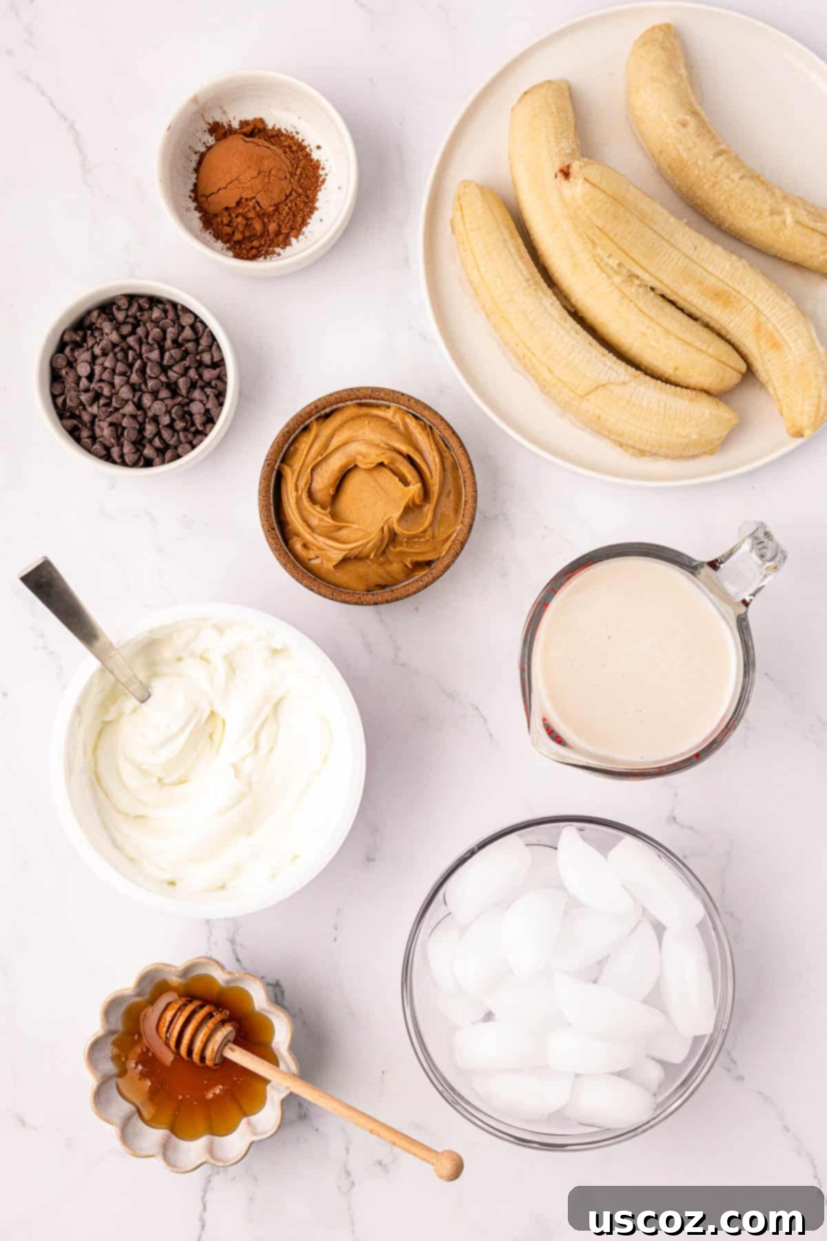Indulgent Chocolate Peanut Butter Banana Smoothie 4 A flat lay photograph of all ingredients needed to make a chocolate peanut butter banana smoothie, including frozen bananas, peanut butter, yogurt, milk, cocoa powder, mini chocolate chips, honey, and ice