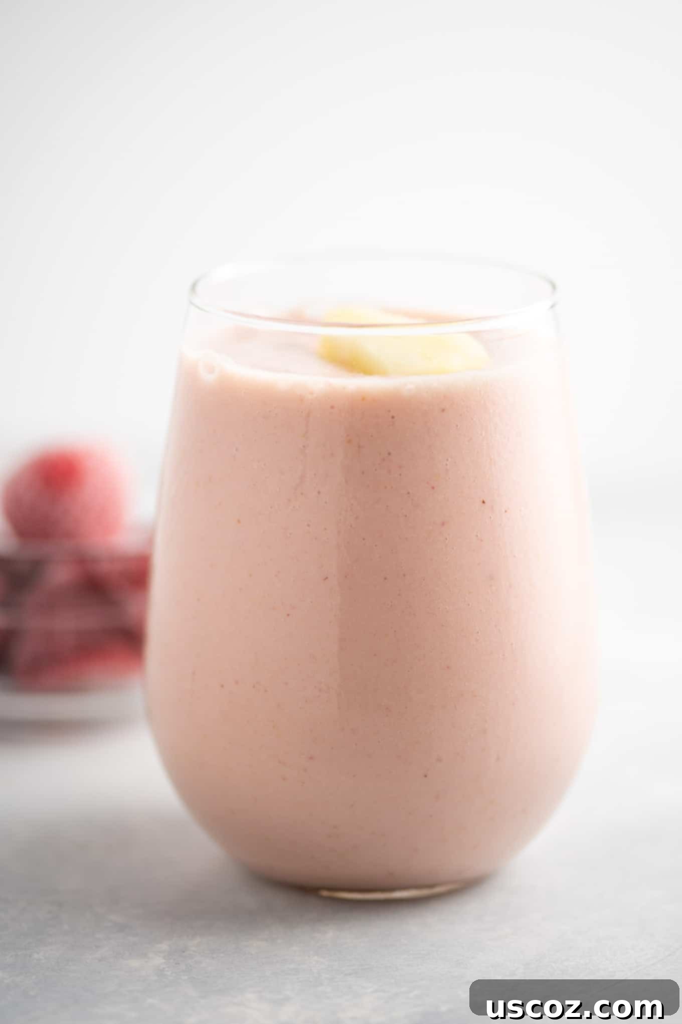 Close-up of a refreshing pineapple strawberry smoothie in a clear glass, with blurred frozen strawberries and pineapple in the background, emphasizing the fresh, cool ingredients.