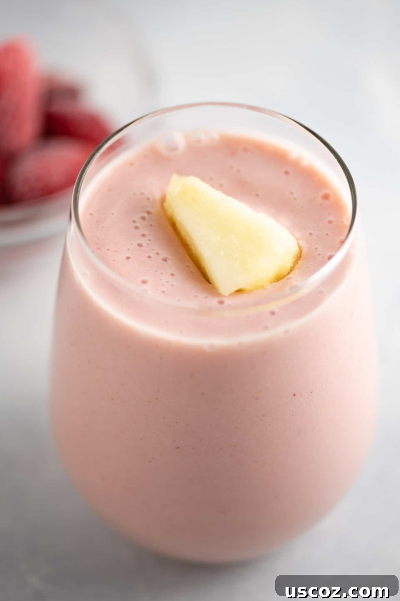 A close-up of a pink strawberry and pineapple smoothie in a glass, with a single frozen pineapple wedge resting on the rim, illustrating a refreshing and healthy drink.