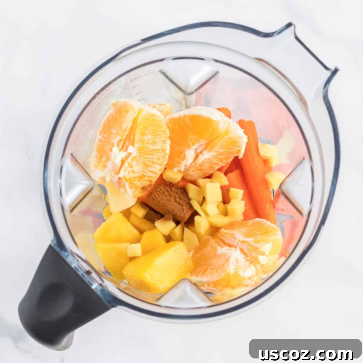 Golden Carrot Orange Smoothie 5 Fresh ingredients for a carrot orange smoothie, including carrots, orange segments, pineapple chunks, ginger, and cinnamon, visible inside a blender jug before blending.