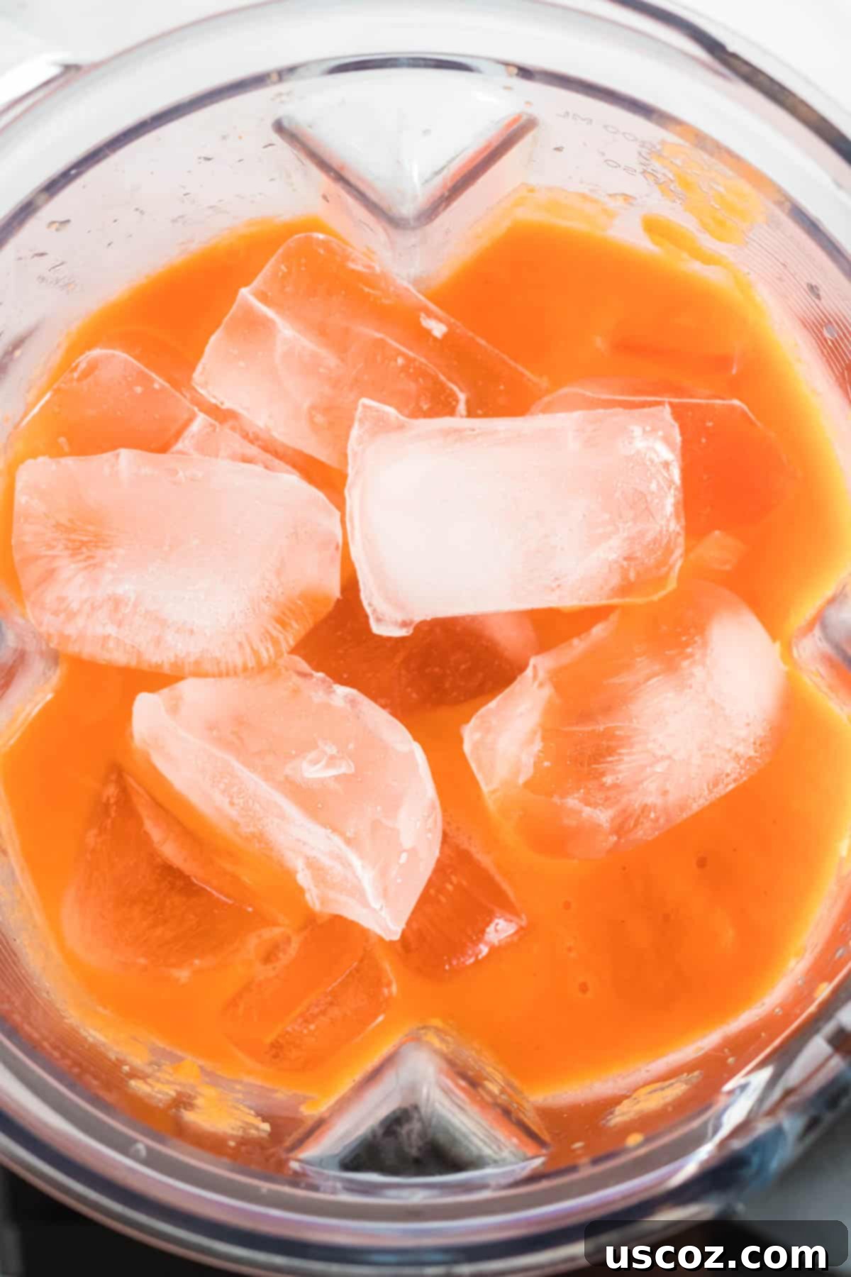 Golden Carrot Orange Smoothie 7 Ice cubes being dropped into a blender filled with a bright orange carrot and fruit smoothie mixture, preparing for the final blend to achieve a frosty consistency.