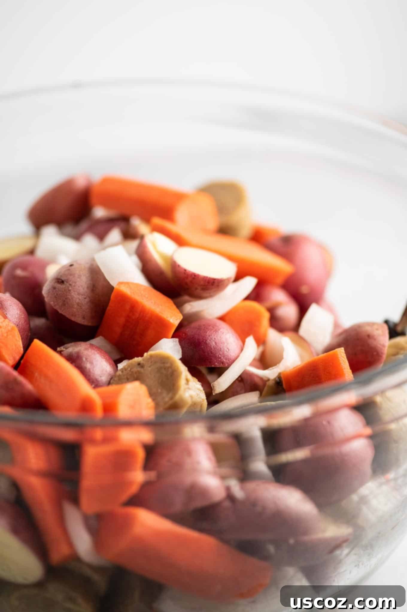 Raw sliced baby red potatoes, carrots, yellow onions, and vegetarian kielbasa in a large mixing bowl, before seasoning.