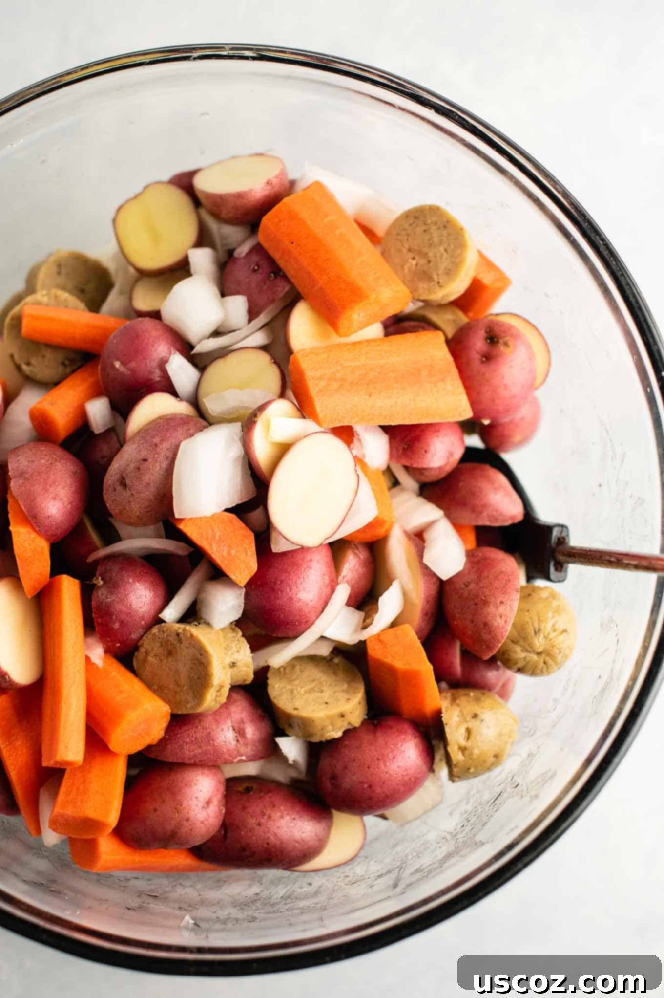 Mixing bowl with sliced vegetarian kielbasa, baby red potatoes, carrots, and yellow onions, tossed with olive oil and spices.