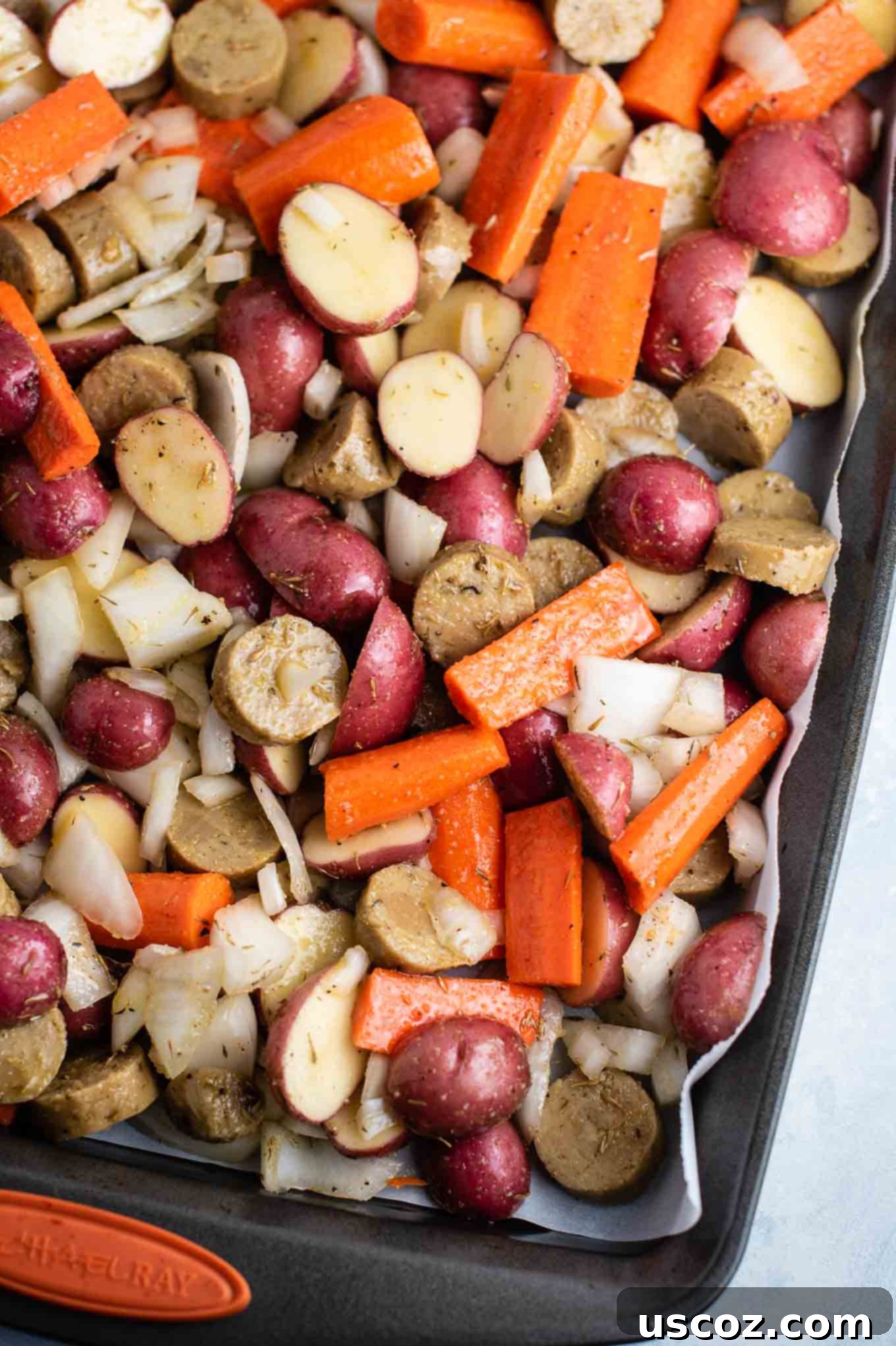 Overhead view of a baking sheet lined with parchment paper, holding mixed raw vegetarian kielbasa and vegetables tossed with seasoning, ready for roasting.