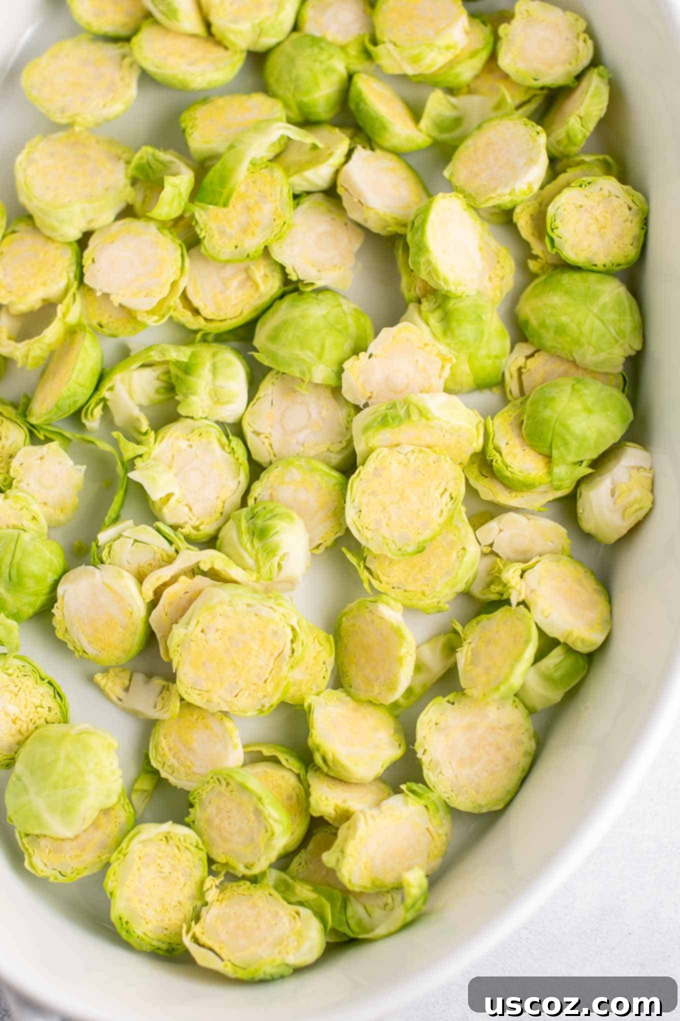 Golden Cheesy Brussels 5 Thinly sliced raw brussels sprouts evenly distributed in an oval white baking dish, ready for roasting.