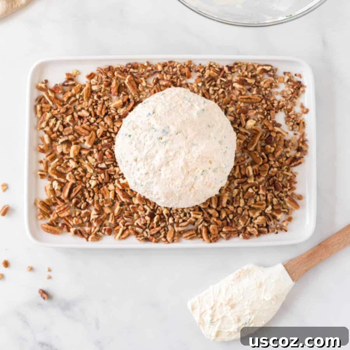 Party Perfect Cheese Ball 9 A freshly shaped cheese ball placed on a plate filled with finely chopped pecan pieces, ready for coating.