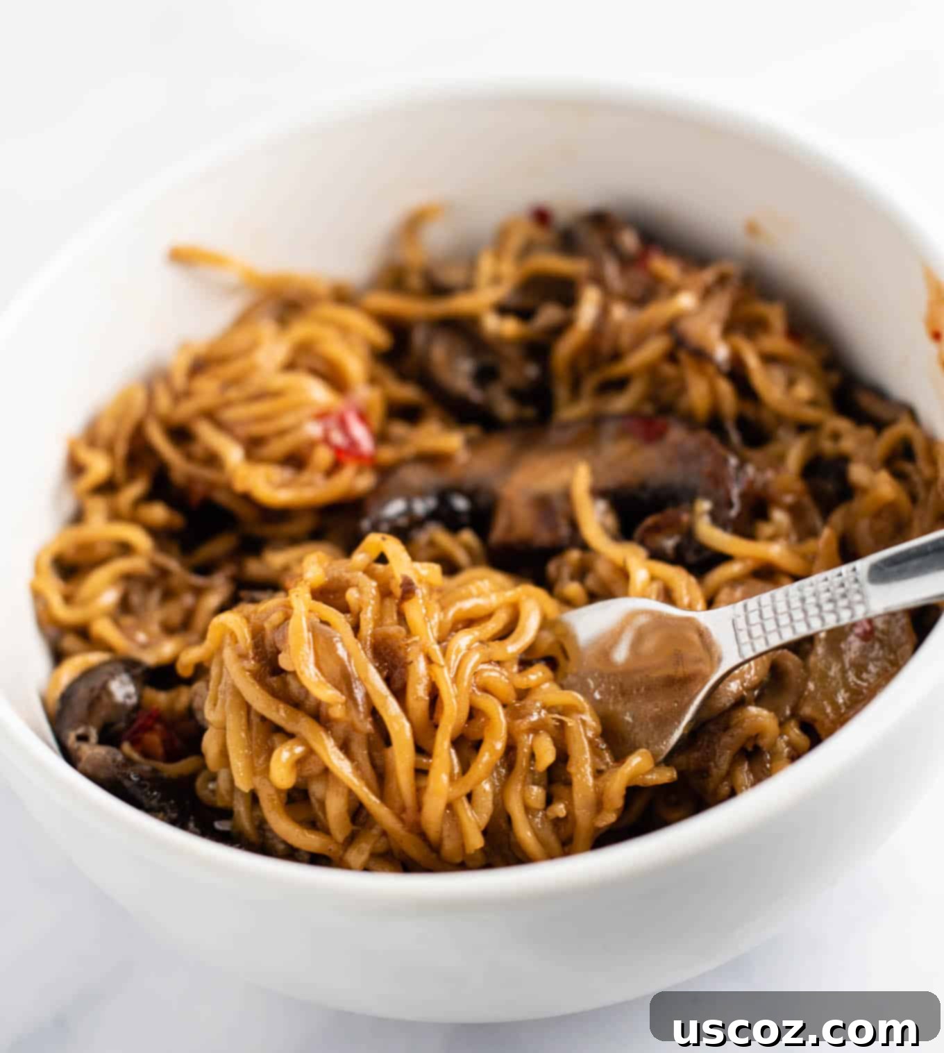 Umami Mushroom Ramen 12 Ramen noodles elegantly twirled on a fork, resting on a bed of stir-fry noodles in a white bowl, inviting a taste.