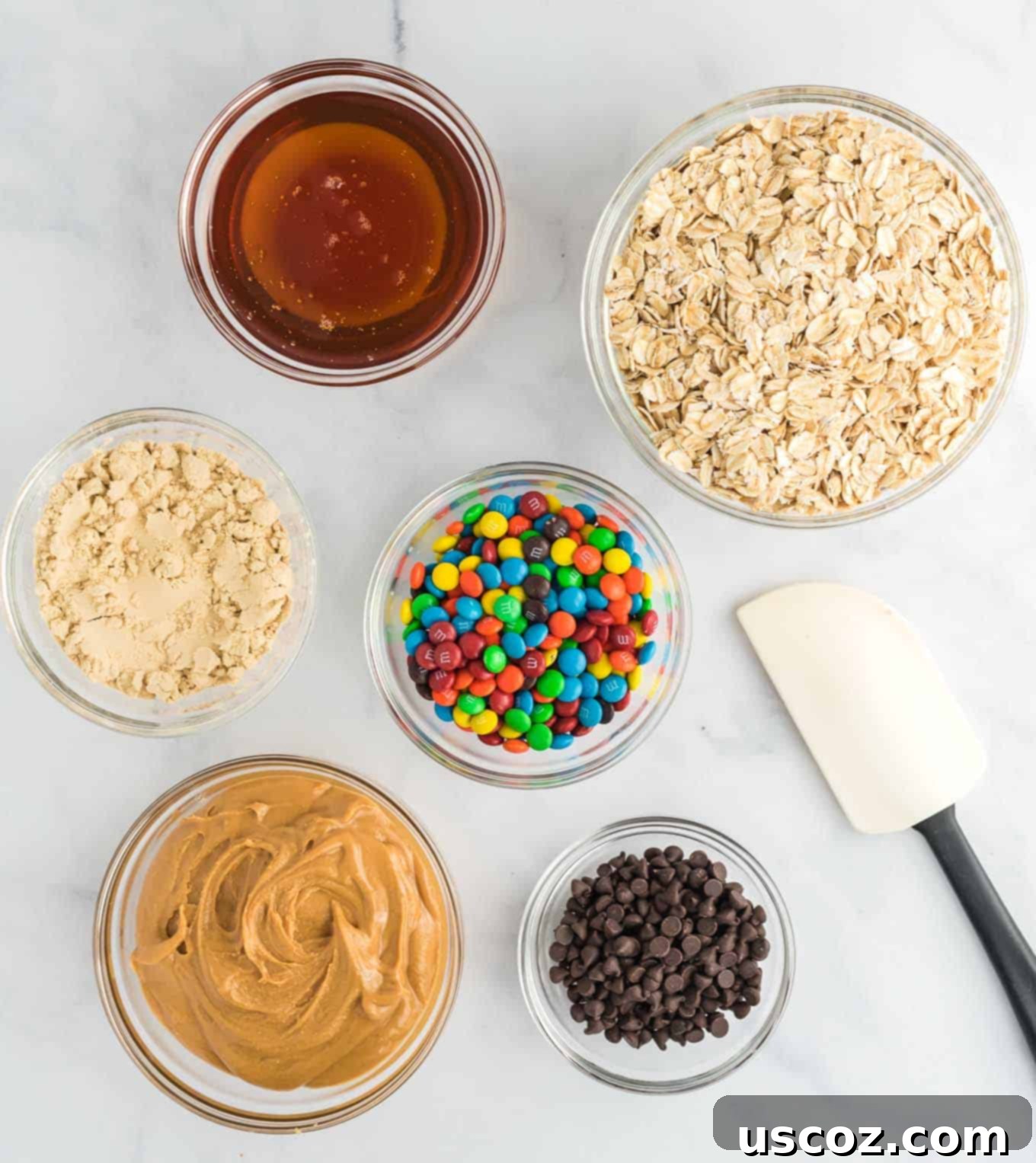 Monster Cookie Energy Bites 4 A colorful display of individual ingredients for Monster Cookie Energy Balls in clear bowls: creamy peanut butter, golden honey, vanilla protein powder, rolled oats, mini chocolate chips, and vibrant mini M&M's.