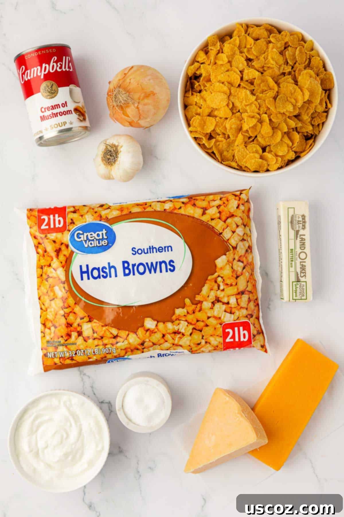 Crispy Cheesy Potato Casserole 4 A flat lay photograph showcasing all the raw ingredients needed to prepare the cheesy potatoes with corn flakes, including frozen hash browns, cheeses, soup, sour cream, butter, onion, garlic, and corn flakes.
