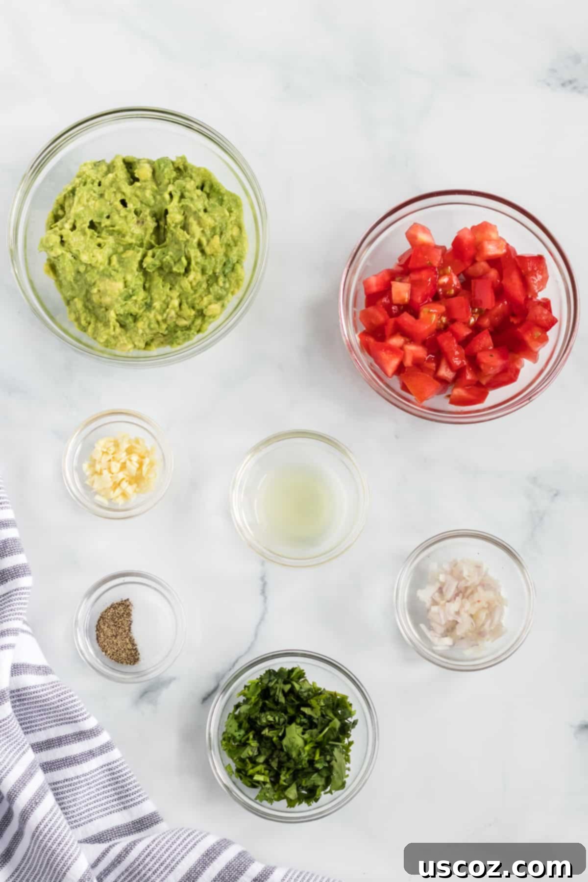 Your New Favorite Guacamole Recipe 3 Individual glass bowls containing fresh ingredients for homemade guacamole: ripe avocados, minced shallots, garlic, chopped cilantro, diced Roma tomatoes, and lime halves.