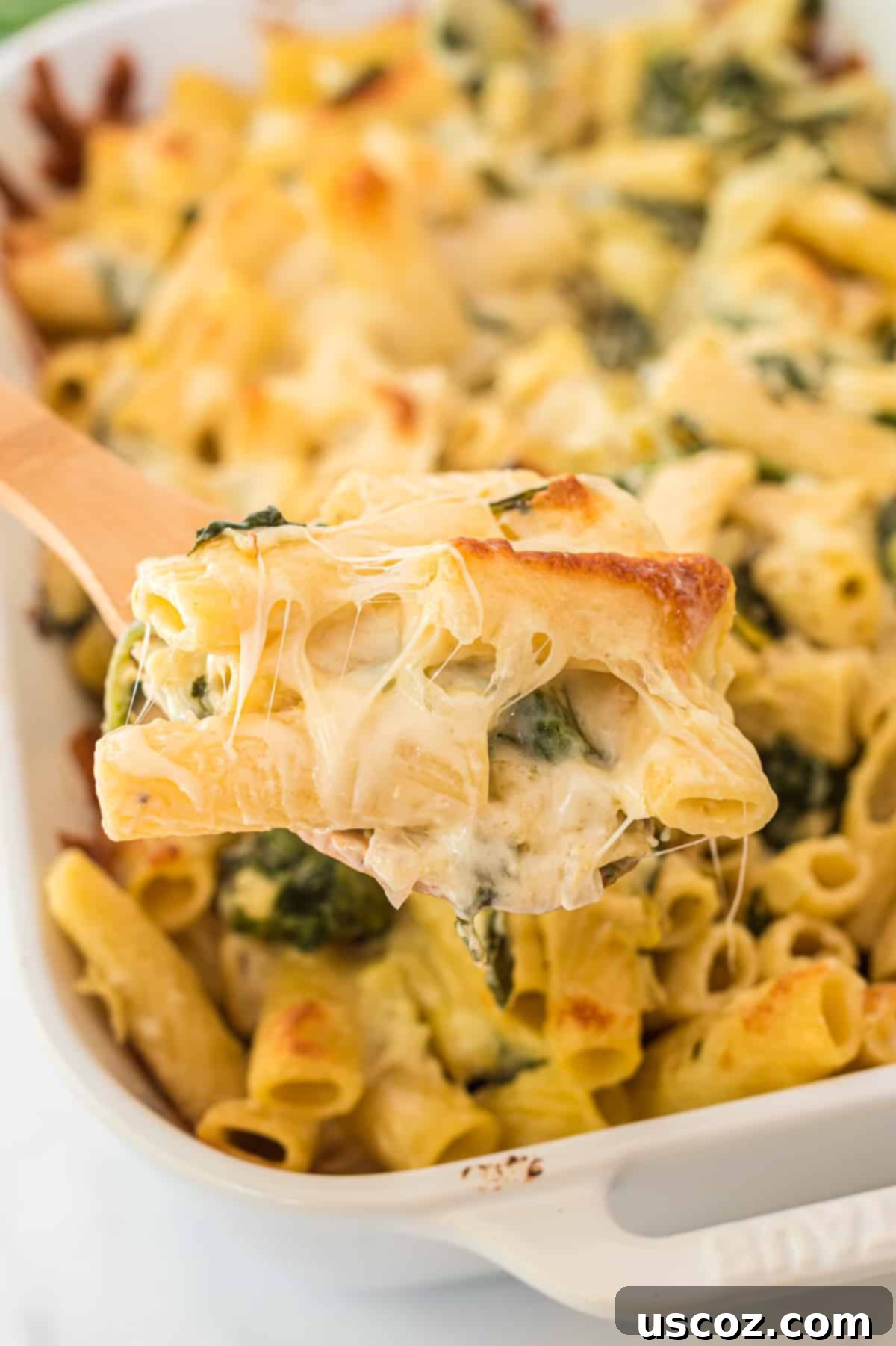 Creamy Spinach Artichoke Pasta Casserole 2 A wooden spoon taking a scoop of creamy spinach artichoke pasta from a white baking dish, showing melted cheese and tender rigatoni