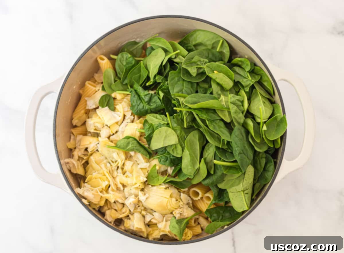 Creamy Spinach Artichoke Pasta Casserole 5 White dutch oven filled with rigatoni pasta, topped with freshly chopped artichokes and spinach, ready to be mixed into the sauce