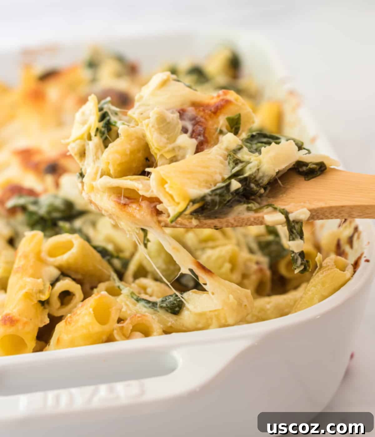 Creamy Spinach Artichoke Pasta Casserole 7 A wooden spoon taking a generous scoop of creamy spinach artichoke pasta from a white baking dish, highlighting the melted cheese and texture