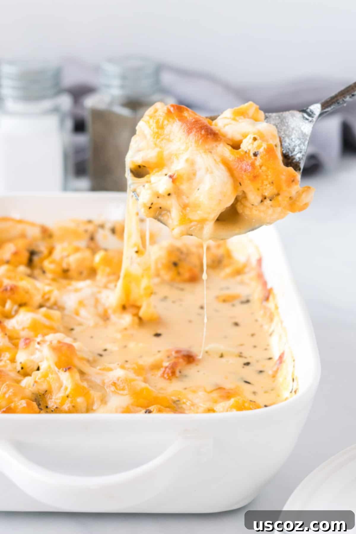 Golden Cheesy Cauliflower 3 A spoon scooping a portion of the cheesy cauliflower casserole from a white baking dish, showing the creamy texture and melted cheese.