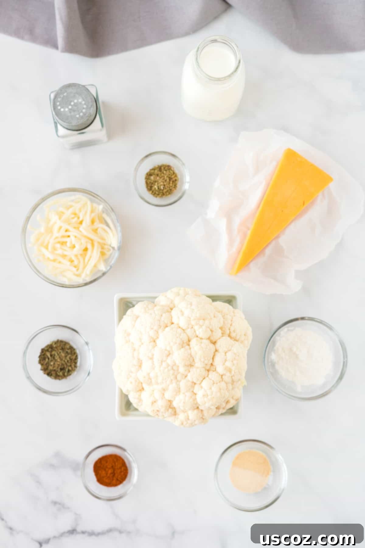 Golden Cheesy Cauliflower 4 A flat lay image showing fresh ingredients to make cheesy cauliflower, including a head of cauliflower, blocks of cheese, milk, and spices, arranged on a wooden background.