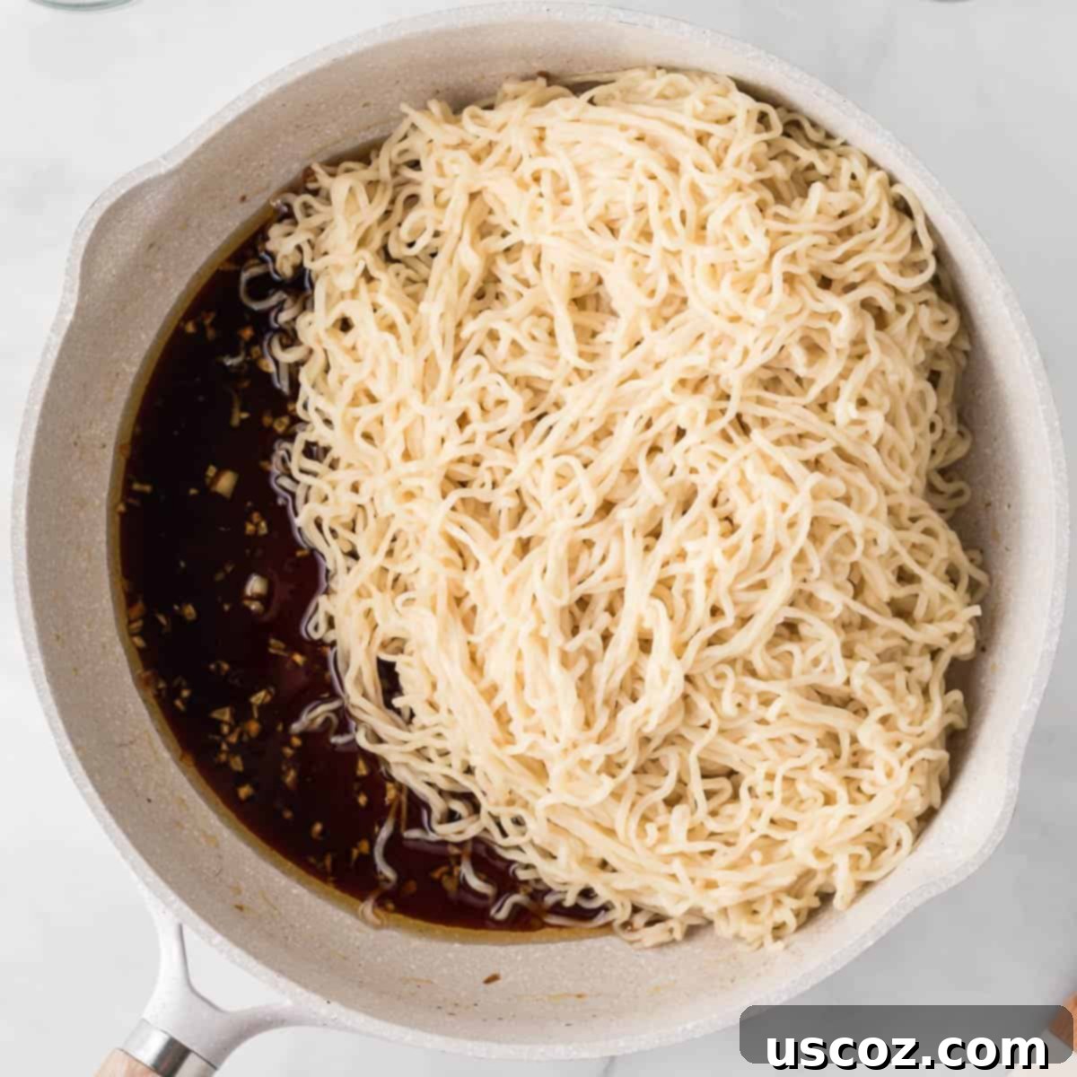 Cooked ramen noodles being added to a skillet filled with the aromatic sesame garlic sauce, ready for a thorough toss to combine flavors.