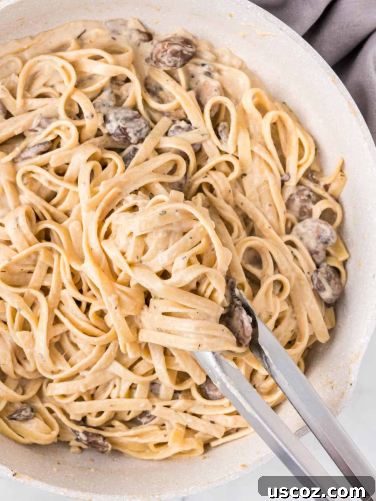 Velvety Mushroom Pasta 13 Tongs picking up a serving of creamy mushroom pasta from a skillet, highlighting the rich sauce and perfectly cooked fettuccine.