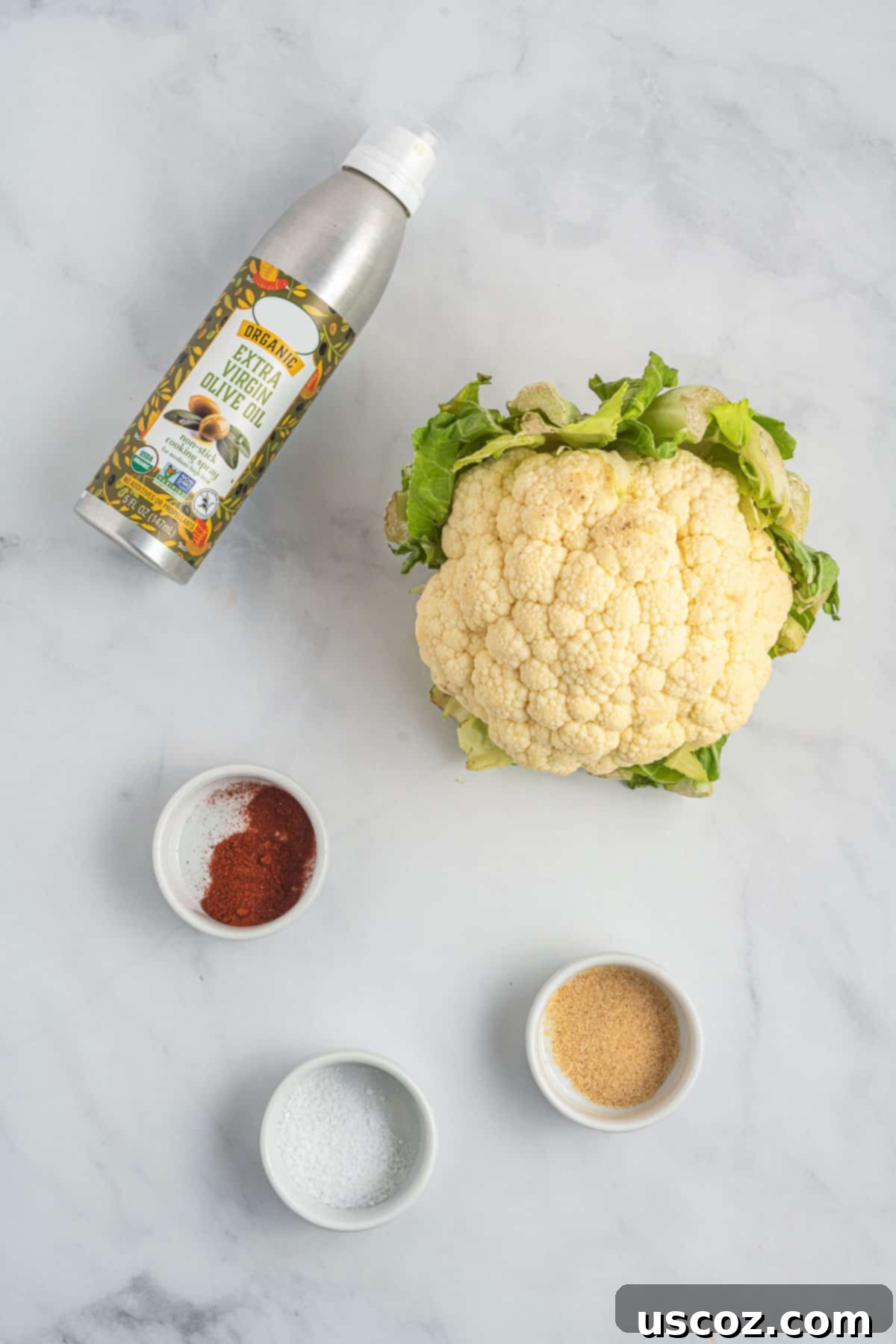 Crispy Air Fryer Cauliflower 3 A fresh head of cauliflower sits beside olive oil spray, salt, garlic powder, and paprika, showcasing the simple ingredients for air fryer cauliflower.