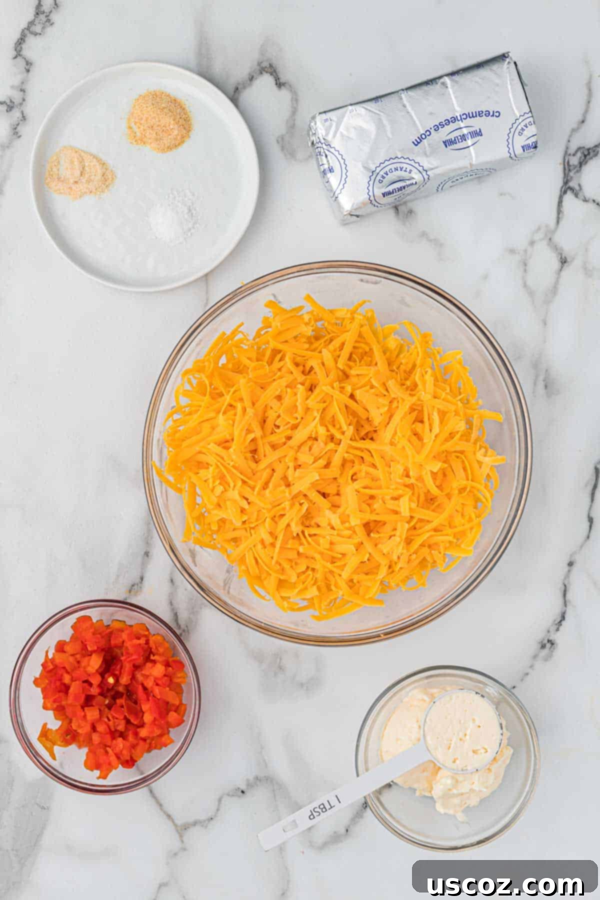 Pimento Cheese 4 All the essential ingredients neatly arranged on a cutting board: a block of sharp cheddar, a package of cream cheese, a jar of diced pimento peppers, mayonnaise, and small bowls of salt, onion powder, and garlic powder.