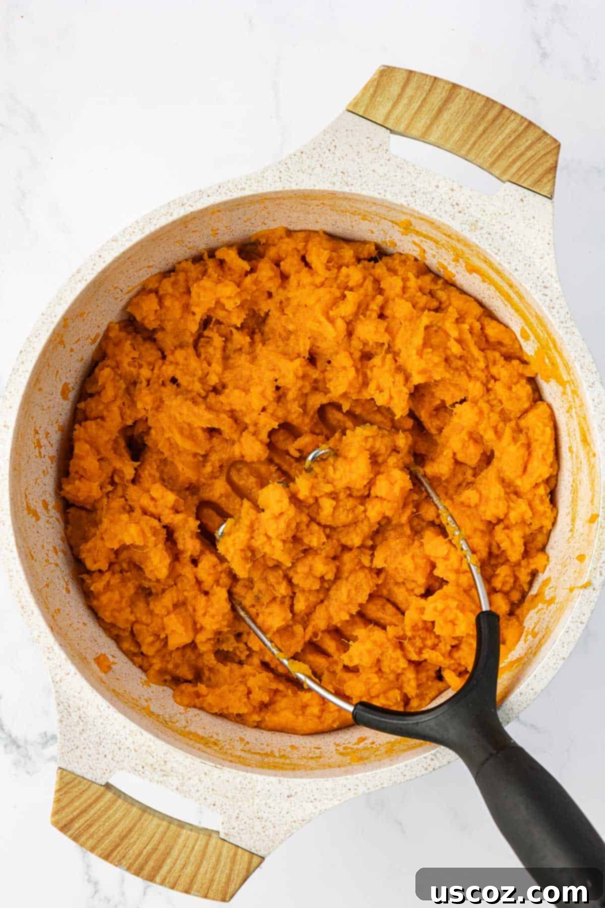 Golden Sweet Potato & Pecan Delight 6 A person actively mashing cooked sweet potatoes in a large pan using a sturdy potato masher, creating a smooth base for the casserole.