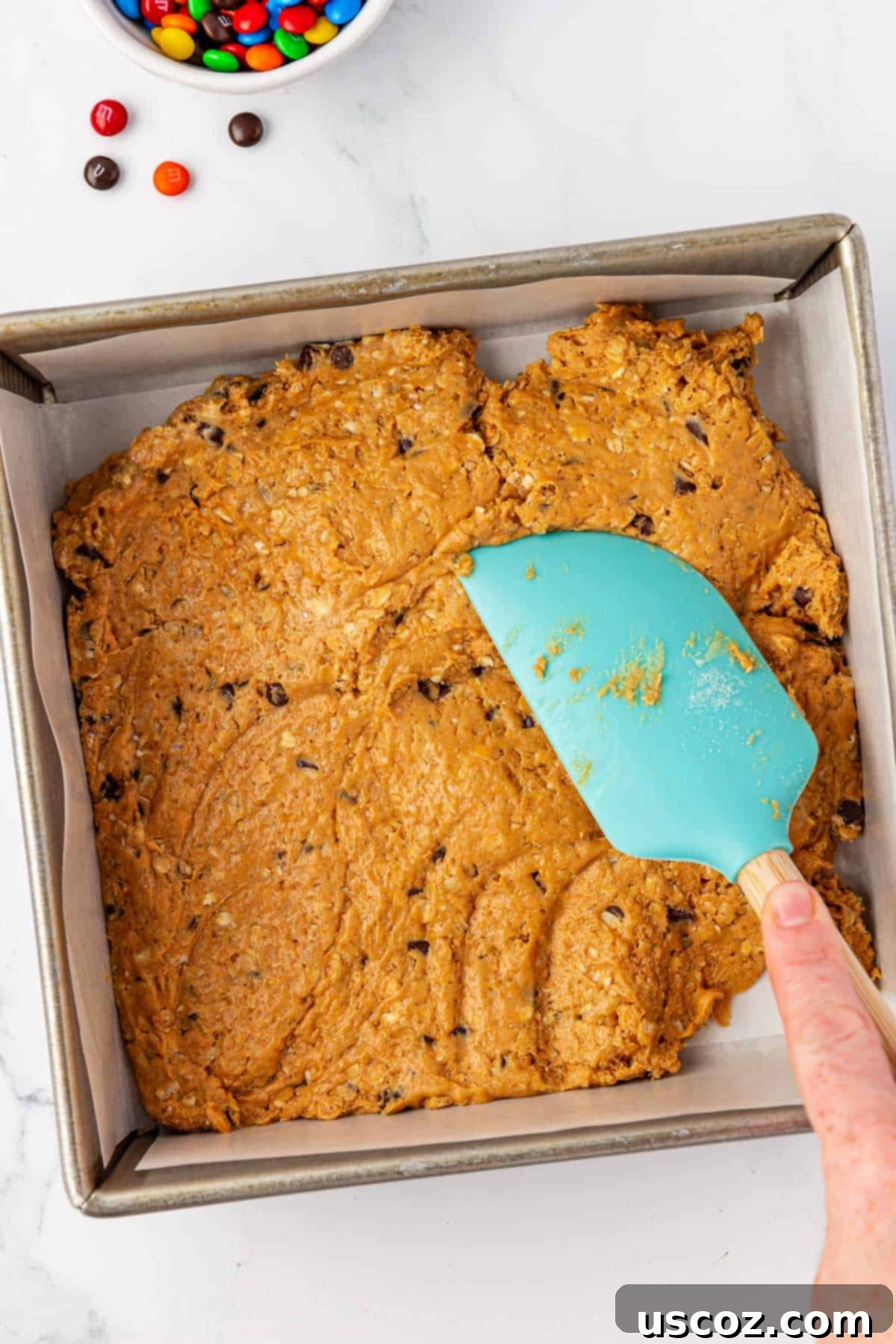 Monster Cookie Protein Bars 7 A rubber spatula pressing the monster cookie protein bar batter evenly into a parchment-lined 8x8 inch baking pan