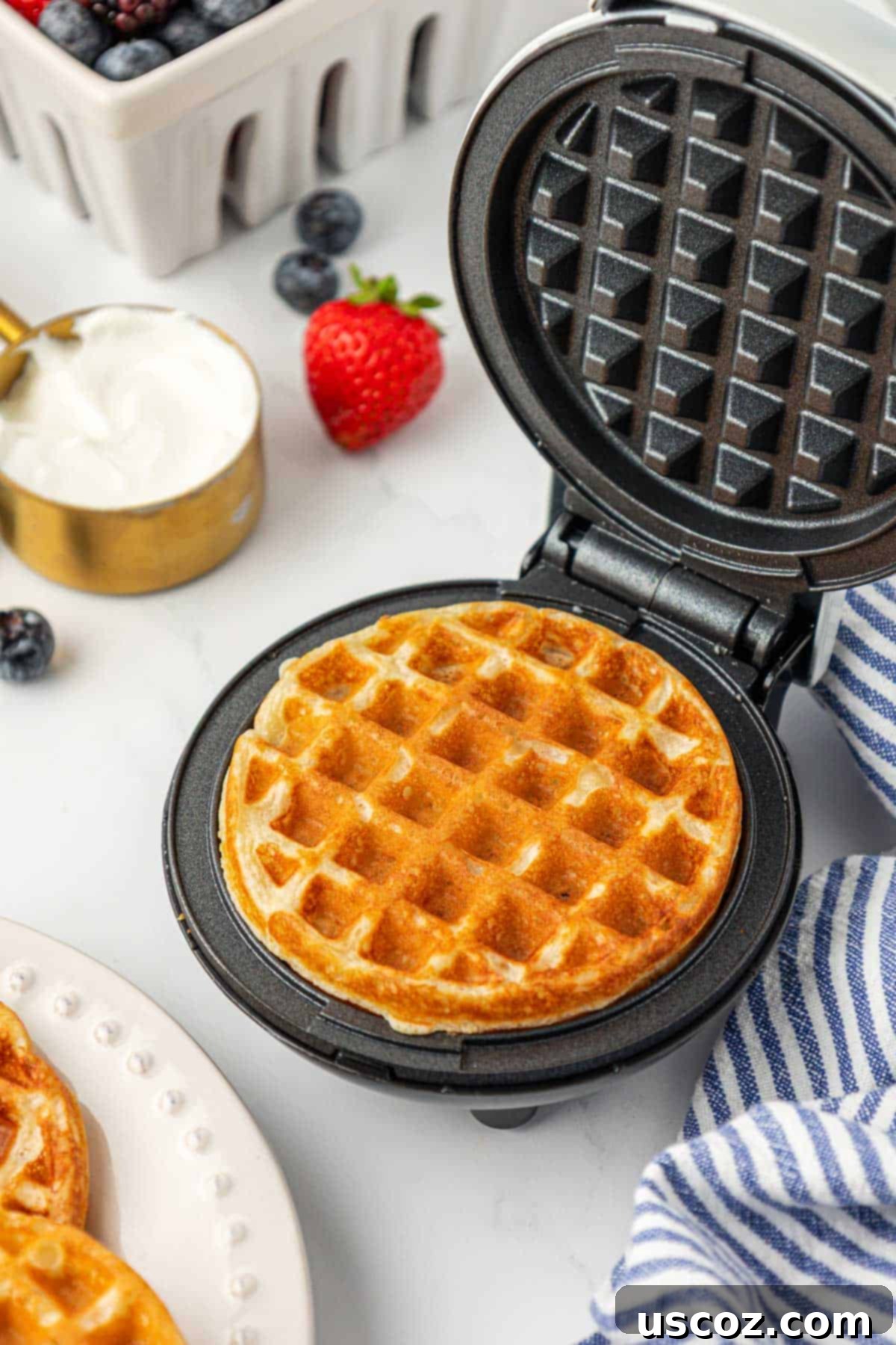 A mini Greek yogurt waffle cooking inside a waffle maker, steam rising, indicating it's nearly done.