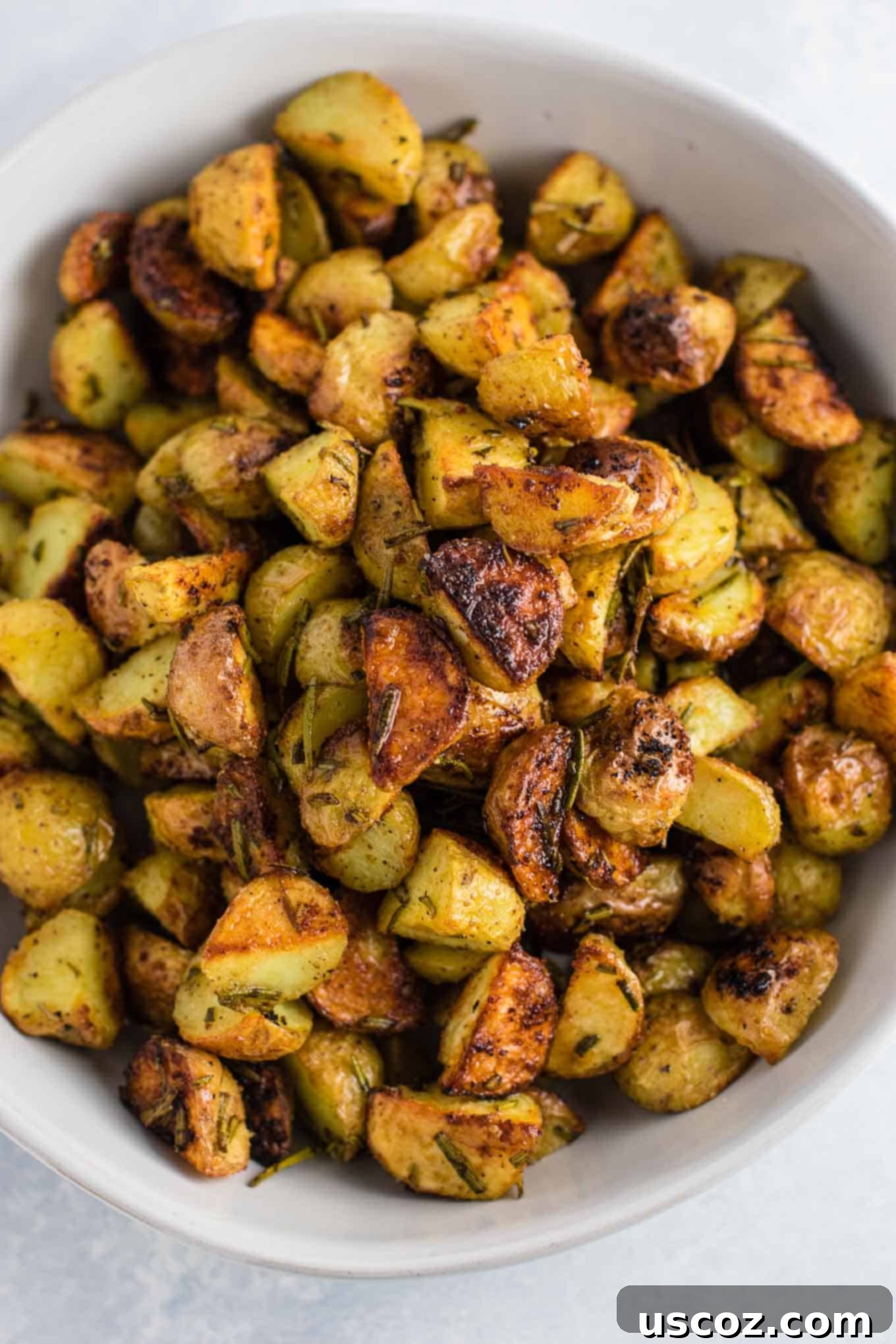A close-up shot of perfectly golden-brown rosemary roasted potatoes garnished with fresh rosemary sprigs in a white serving bowl.